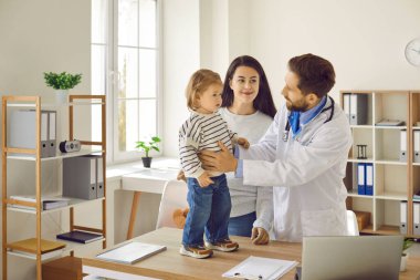 Caring, friendly doctor examines his little patient, smiling mother at the next consultation of her child with pediatrician in the office of modern medical institution. Healthcare concept.