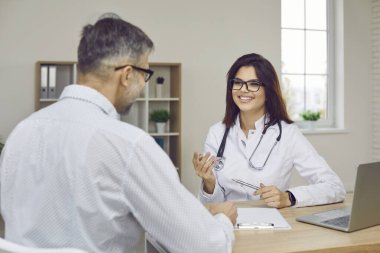 Modern medicine. Friendly caring female doctor talking to patient during medical consultation in hospital. Smiling young woman in medical gown sits at table and explains something to middle-aged man.