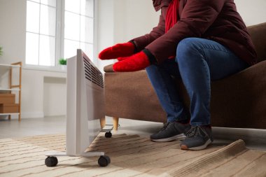 Young man is sitting at home on the sofa in warm jacket and knitted red mittens near heater, freezing from the cold. There is no heat in house, there are problems with heating. Energy supply concept.