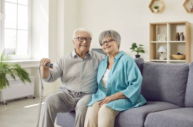 Evdeki oturma odasında koltuk değneği olan mutlu bir son sınıf aile eşi ve koca portresi. İki gülümseyen gri saçlı emekli adam ve kadın emekliliğin tadını çıkarıyorlar..