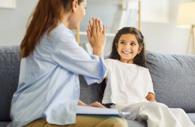 Child girl and counselor celebrate successful therapy session support. Psychologist with clipboard high fives kid in clinic office during consultation. Family psychotherapy and mental health.