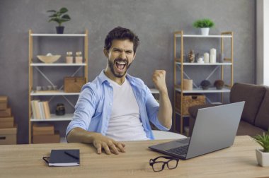 Hurray. Euphoric handsome business man fist pumping sitting at work table with laptop computer. Happy emotional guy feeling excited as he finds great deal, finishes freelance project, gets job done