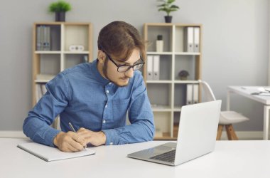 Concentrated young man with glasses makes notes while working with laptop at home. Businessman or student jotting down information from internet or important things during distance business training.