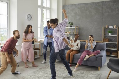 Happy positive diverse college students and friends dancing and having fun at a party at home. Mixed race group of cheerful people watching young man dance, applauding and enjoying good time together
