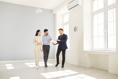 Professional realtor presenting new apartment to young family couple and discussing terms of purchase contract. Real estate agent showing clients new home or office space, talking about its benefits.
