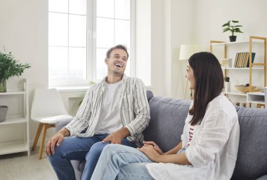Cheerful young family couple sitting on comfortable sofa in living room and laughing together. Happy loving husband and wife having fun, telling jokes, enjoying home leisure time during weekends.