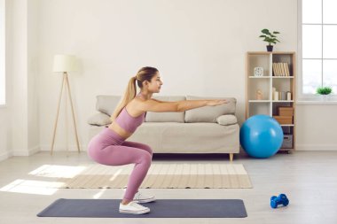 Fit young woman in sportswear do squats train exercise at home. Millennial girl workout do sports in living room, follow healthy lifestyle. Female coach or trainer working out on lockdown indoors.