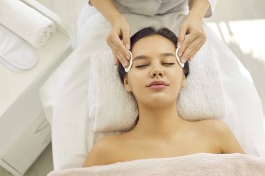 Happy beautiful woman lying on spa massage bed in beauty parlor and enjoying modern facial treatment. Beautician removing makeup with cotton discs and cleaning skin before rejuvenating detox procedure