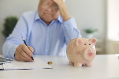 Domuz kumbarası bir masanın ortasında duruyor. Not defterine not alan yorgun ve ciddi bir kıdemli adamın yanında. Emekli bir emeklilik planı ya da aylık ödeme hakkında düşünür ve mali işler yönetir.