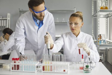 Laboratuvarda çalışırken kan örneklerinin analizi üzerinde çalışan iki bilim adamı. Erkek bilim adamı, birkaç test tüpünden birine sıvı eklemek için pipet kullanan bir meslektaşını gözlemliyor..