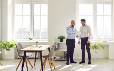Business people working in the office. Two young men discussing some professional matters while standing in a modern spacious office workplace lit with bright sunlight