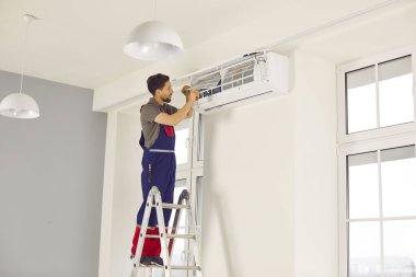 AC service technician installing new air conditioner. Man in uniform workwear overalls standing on step ladder and fixing air conditioner on white wall. Air conditioning installation at home concept