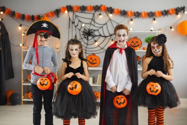 Group of children in different Halloween costumes. Bunch of kids dressed up as spooky characters standing in room decorated with black cobwebs and orange pumpkins and holding trick or treat buckets