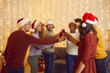 Good friends are multiracial men and women, drinking wine and clinking glasses at New Years Eve or Christmas party. People in Santa hats have fun at home in living room with garlands on walls.