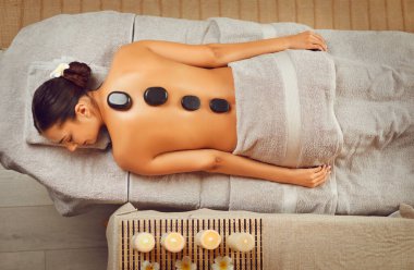 Serene young woman having hot stone massage in spa salon. View from above of relaxed attractive girl lying on her stomach with basalt stones along spine. Beauty treatment, body care concept