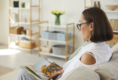 Nostalgia. Woman looks at her family photo album and remembers wonderful moments in her life. Close up side view of woman sitting on sofa at home in living room. Concept of family history and memories