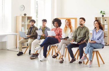 Team of diverse people with paper notes attending corporate business seminar meeting in office, sitting on chairs as audience members, listening to speaker, waiting for their turn to make presentation