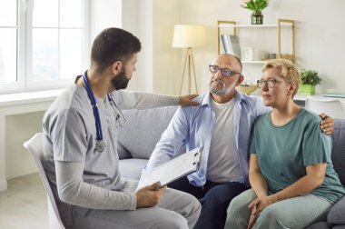 Male healthcare worker and nurse comforts a senior elderly couple during a home visit, assessing wellbeing and offering medical support in livingroom. Nursing service and medicine comcept. 