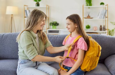 Mother and daughter start school preparation morning. At home, mom adjusts a backpack strap, offering support before class in a family livingroom with daylight. Kind help, education comcept.