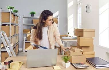 Woman entrepreneur is working at a warehouse, conducting inventory of packages using a laptop. She is focused and surrounded by boxes and parcels, managing the storage efficiently.