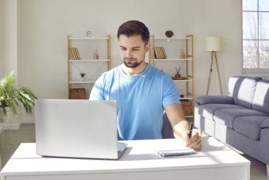 Genç ve çekici bir iş adamının portresi ofiste ya da evde dizüstü bilgisayarda çalışıyor ve masada yazı yazıyor, notlar alıyor, hesaplamalar yapıyor. Uzak ve serbest çalışma konsepti.