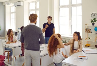 Bearded teacher communicates with student through desk against background of large bright window in modern classroom. Classmates listen carefully to their conversation. Quality education in America.