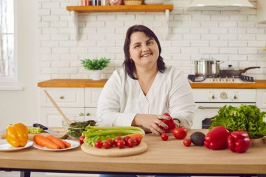 Portrait of happy fat overweight young woman looking at the camera and smiling standing in the kitchen with a lot of fresh vegetables at home. Diet restrictions, healthy diet, and weigh loss concept.