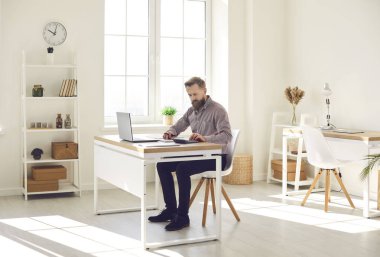 Serious businessman working in cosy, sunny office interior. Busy man doing paperwork, using calculator and checking some documents while sitting at desk with modern laptop computer