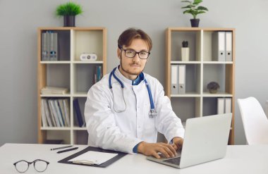 Profile portrait of confident young male doctor sitting at laptop at his workplace. Man in medical clothes is sitting at table in hospital or private clinic and looking at camera. Healthcare concept.