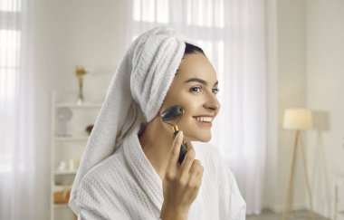 Happy young woman with bath towel turban on head massaging her pretty face with jade facial roller to stimulate lymphatic drainage, increase blood flow, depuff and have better looking, radiant skin