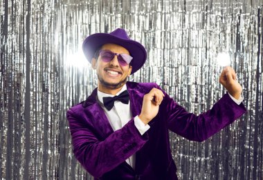 Happy funny young black man wearing tacky unfashionable purple velvet suit, hat and sunglasses dancing at party. Goofy ethnic guy dancing on concert show stage with shiny foil fringe studio background