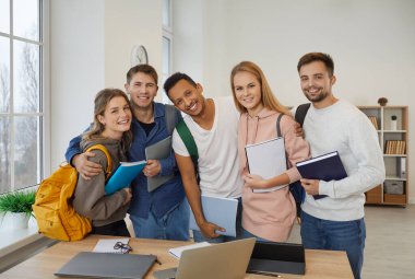 Diverse team of five happy university students. Indoor group portrait of smiling mixed race multiethnic friends and interracial couples with books and backpacks standing together and looking at camera