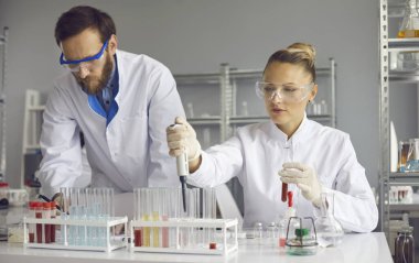 Team of focused scientists working together on sample analyzes while working in a lab. Male scientist makes notes while his female assistant uses a pipette to add liquid to one of several test tubes.