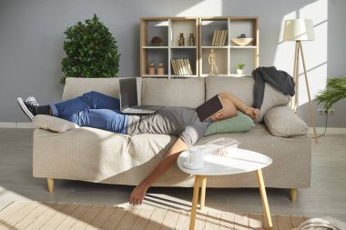 Lazy university student falls asleep while studying at home. Tired young man lying on sofa in living room interior, with book on face, laptop computer on tummy and messy food leftovers on coffee table