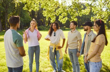 Company of happy young friends talking with each other standing in a circle in summer park. Smiling men and women students in casual communicating gathering in nature outdoors. Friendship concept.