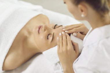 Kadın masör güzellik salonunda ya da kozmetoloji dolabında gözleri kapalı yatan genç bir kıza yüz masajı yapıyor. Profesyonel kozmetik uzmanı kaplıcadaki kadına masaj yapıyor..