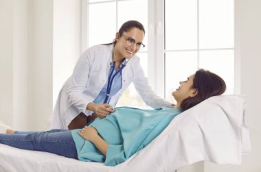 Friendly smiling gynecologist examining heartbeat in the abdomen of pregnant young woman lying on the couch in medical clinic. Obstetrician doctor or nurse checking her patient expecting a baby.