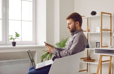 Young confident bearded business man with laptop working using mobile phone, sitting in the chair with modern computer at office or at home. Working at home, freelance work concept.