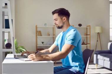 Side view portrait of a young attractive business man working on a laptop at the office or at home and writing at the desk making notes, calculations. Remote and freelance work concept.