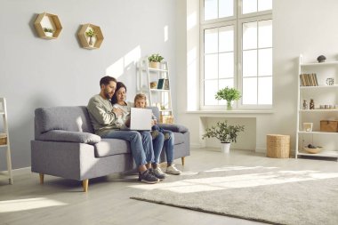 Happy family spend time in spacious room at home. Couple with child watch online movie on laptop sitting on sofa in cozy light living place space with white grey walls, shelves, house plants and wifi
