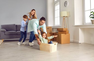 Happy parents and kids having fun riding in living room enjoying relocation day. Dad, mom and son pushing cute little daughter sitting inside of cardboard box. Loan mortgage, housing improvement