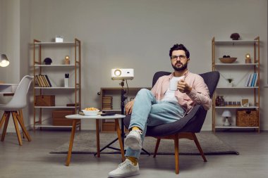 Relaxed young man sitting in a comfortable armchair in the living room at home, using a modern video projector, watching a movie, having a snack, and drinking tea