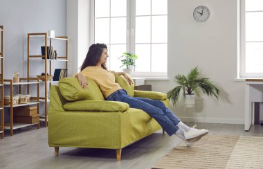 Happy, relaxed woman sitting on a comfortable sofa at home. Beautiful young girl relaxing on a comfortable green couch in a modern living room interior in her apartment