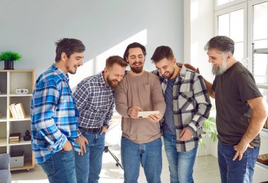 Overjoyed smiling male friends watching photos on smartphone and having fun together. Group of happy mature men standing in the living room at home enjoying funny videos on mobile phone.