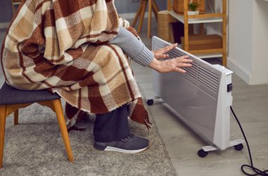 Senior man using an electric heater in winter. Cropped shot of an elderly man in a plaid blanket sitting in a cold room at home, freezing and warming up his hands by a modern electric heater