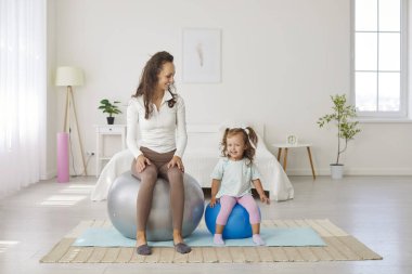 Mother and her young daughter are engaged in playful fitness activities on colorful fitballs. Mother sits on larger fitball, smiling at her daughter, who is excitedly balancing on smaller fitball.