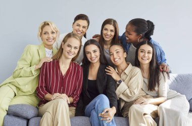 Happy smiling multiracial women team sitting on sofa, looking at camera and making group photo. Successful confident joyful female colleagues or friends in casual wear hugging and laughing together.