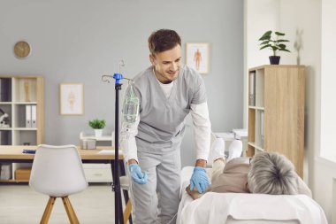 Young friendly nurse man or caregiver help sick senior elderly woman patient in receiving IV drip infusion and vitamin injection therapy lying on couch in medical clinic. Health care concept.