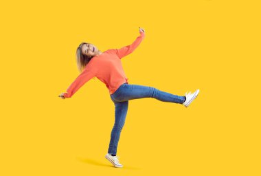Studio shot of funny smiley young girl in comfortable casual clothes having fun and fooling around. Happy pretty woman in orange sweatshirt and blue jeans walking isolated on vibrant yellow background