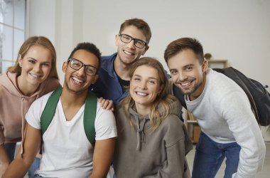 Diverse team of five happy, cheerful college or university students, classmates and friends. Indoor group portrait of multiracial young people with backpacks looking at the camera and smiling